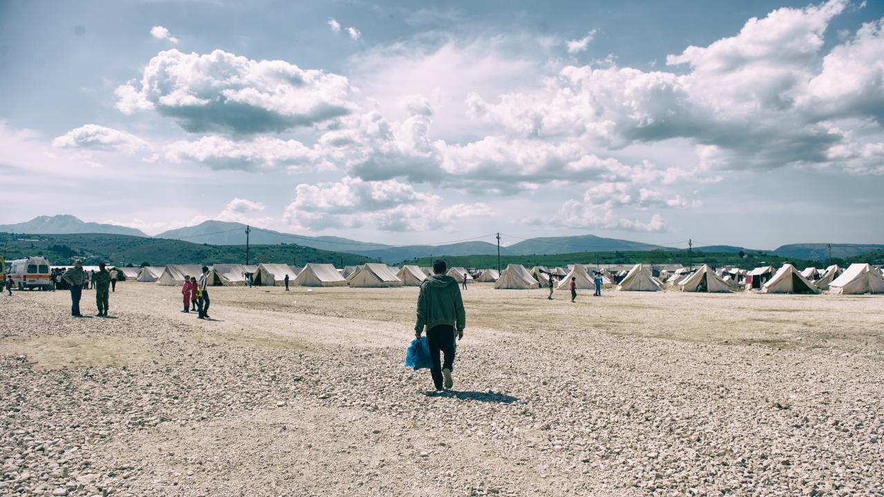 A man carrying a plastic bag in one hand, walking on gravel towards a refugee camp