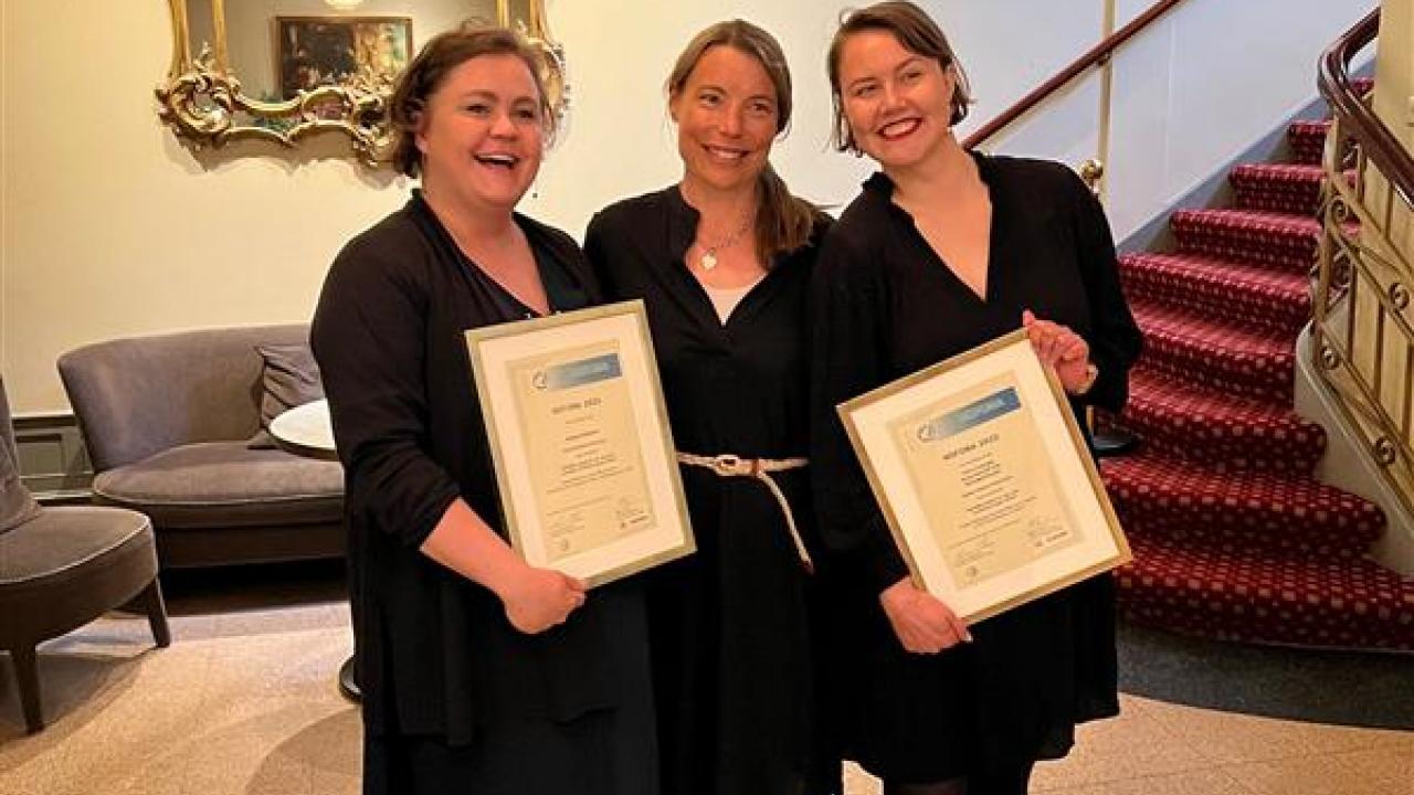 Three brunet women standing close to each other holding two framed certificates of the prices won during the conference. All women are wearing classy black dresses.
