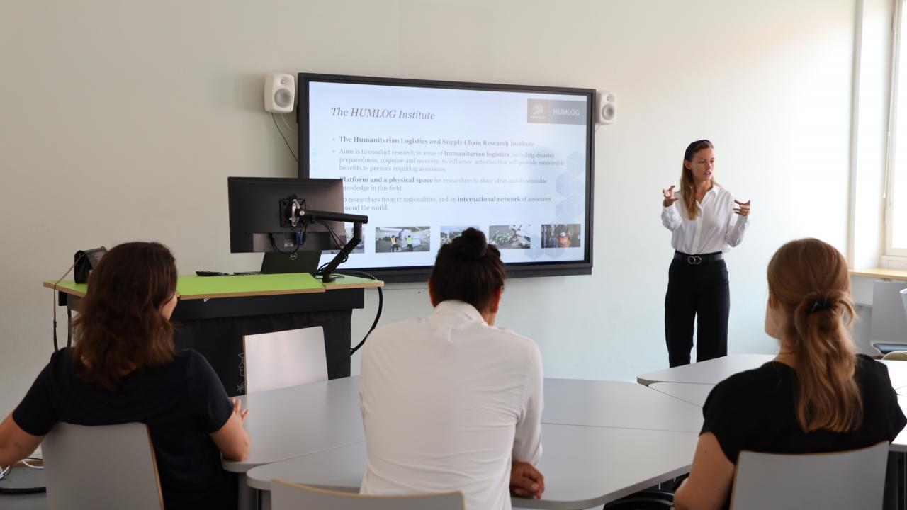 A picture of a brightly lit classrom with a large screen on the back wall with a young woman teaching wearing a white shirt and black pants. From the audience three people's back profiles are visible.