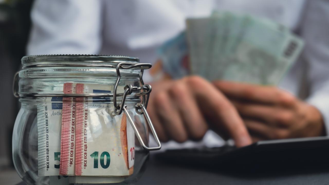 Money in a jar and a person writing on a computer holding more money