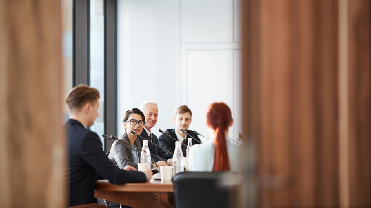Men and women sitting at a conference room table with mics, discussing with each other.