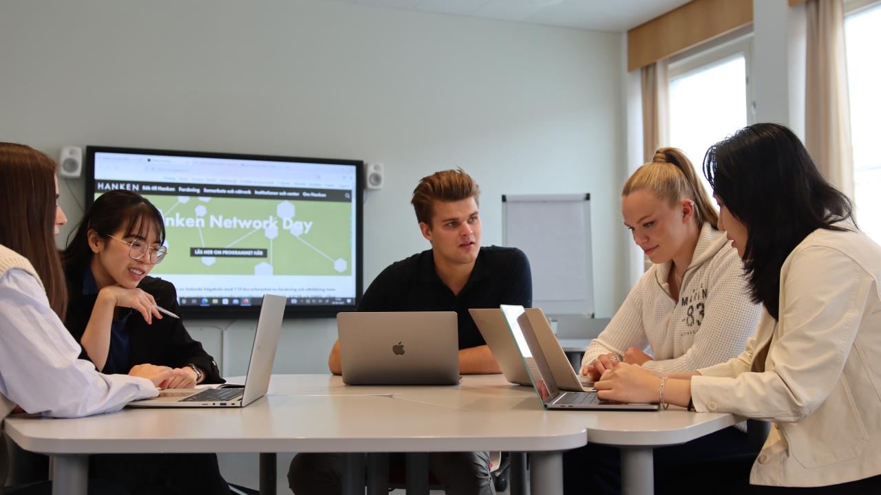 Några studerande sitter vid ett bord och jobbar på sina datorer.