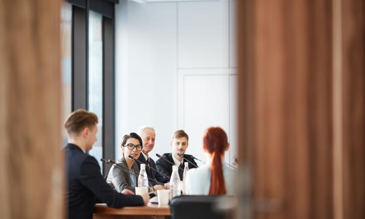 Men and women sitting at a conference room table with mics, discussing with each other.