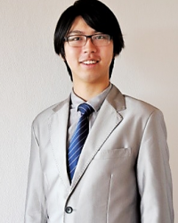 A man with black hair wearing glasses. The man in his late 20's is wearing a silver suit with a blue tie.