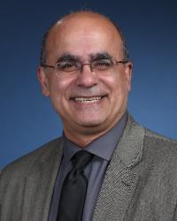 A professional picture of a elderly man wearing a grey blazer, black tie and dark grey shirt, Joe's smiling at the camera and wears glasses.
