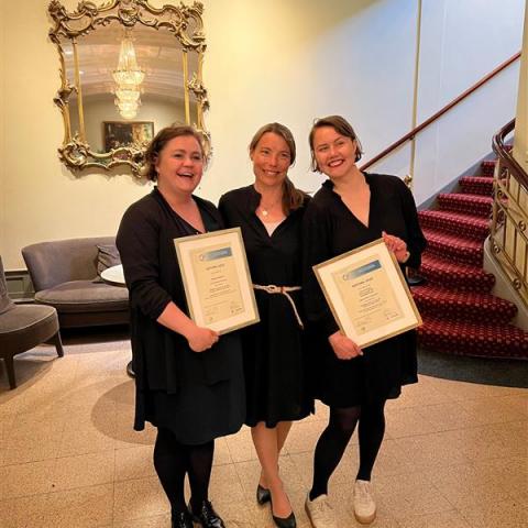 Three brunet women standing close to each other holding two framed certificates of the prices won during the conference. All women are wearing classy black dresses.