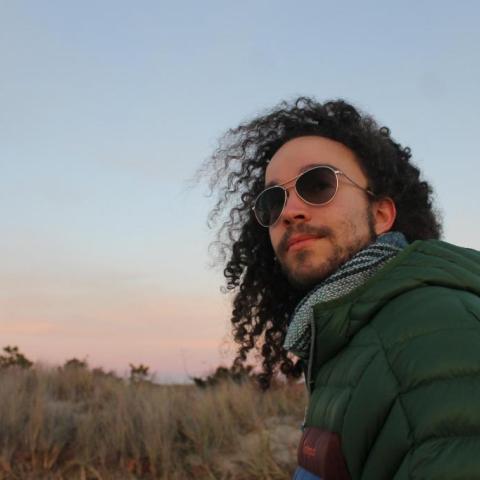 Kieto with sunglasses and jacket and scarf with sky behind on the beach